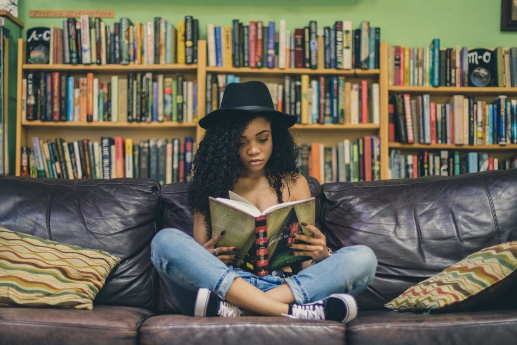 black woman reading a book on a brown sofa seating indian style in front of a bookshelf